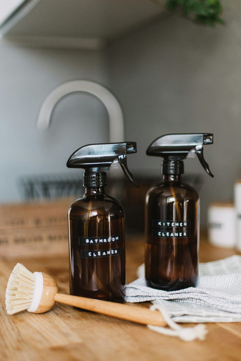 Two brown spray bottles of cleaner on wooden counter next to cloth and wooden brush