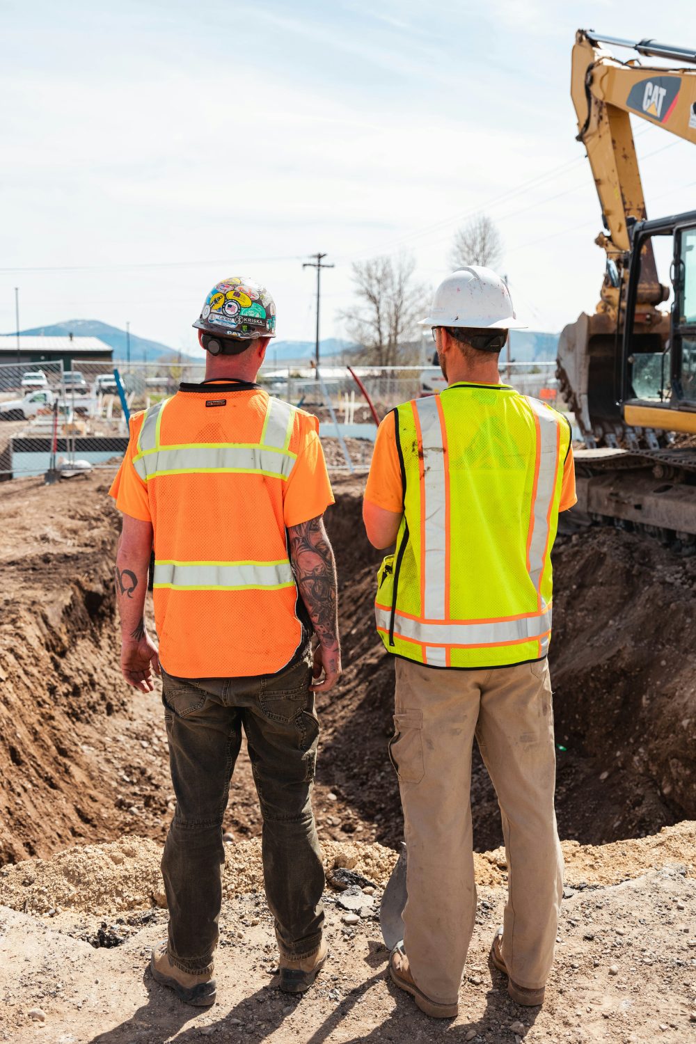 Two male construction workers in high-vis jackets and hard hats standing on edge of earth pit next to yellow excavator 