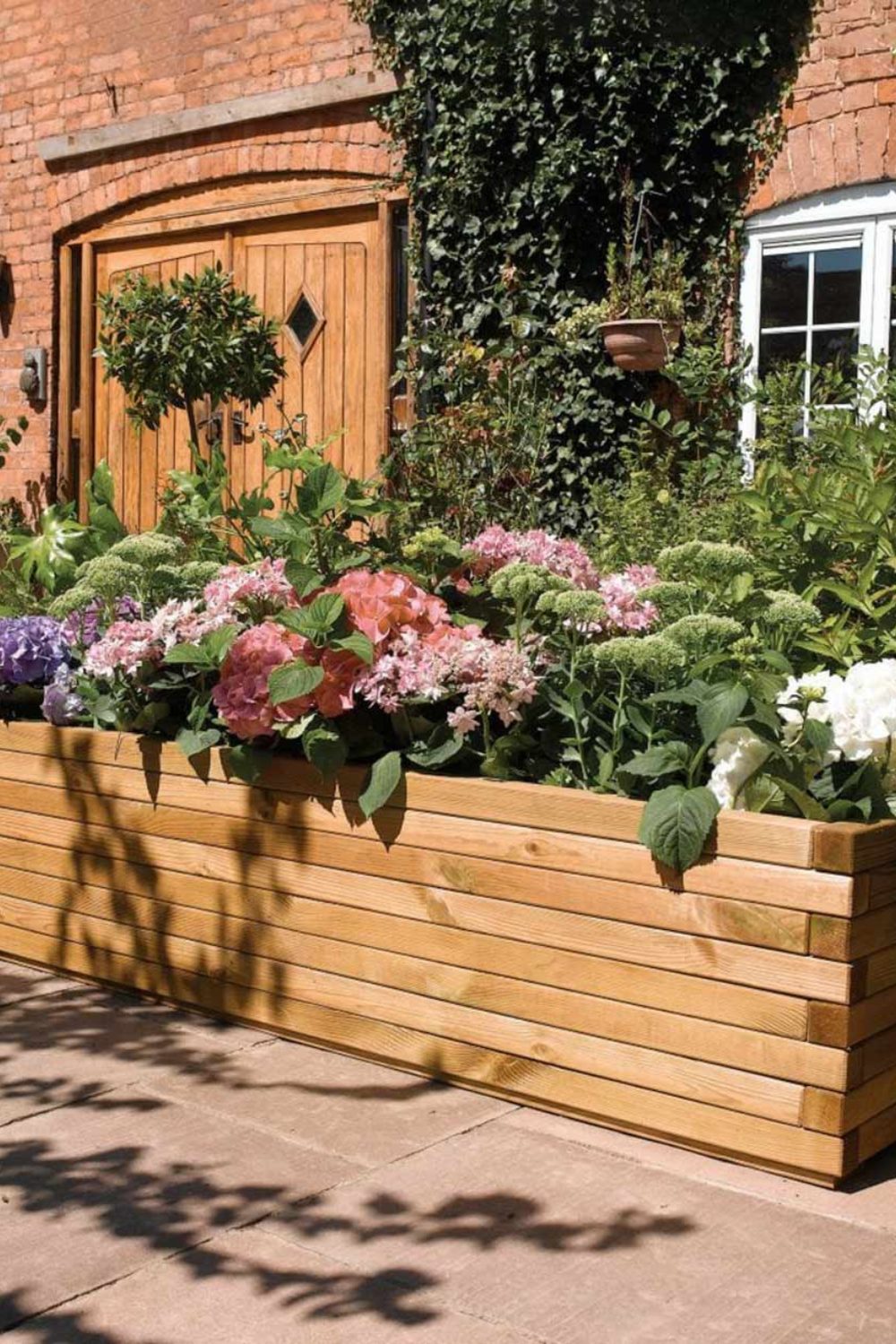 Wooden planter holding pink flowers in front of a red brick house covered in ivy with wooden double doors