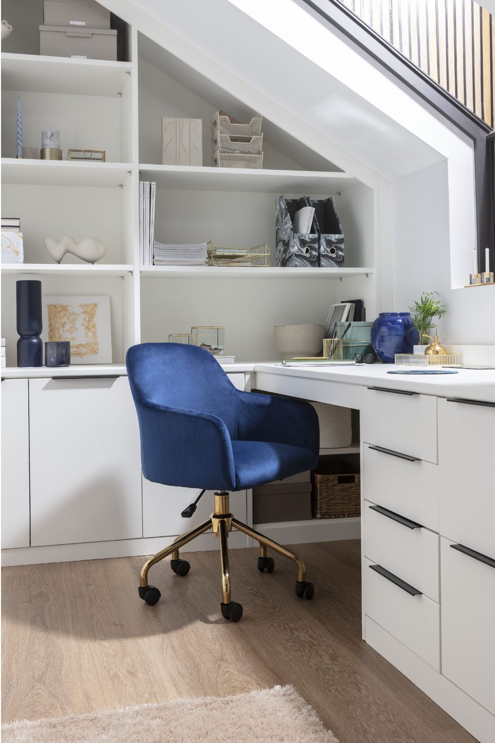 Home office in loft space with blue chair, white cupboards and white shelving holding files, vases and papers