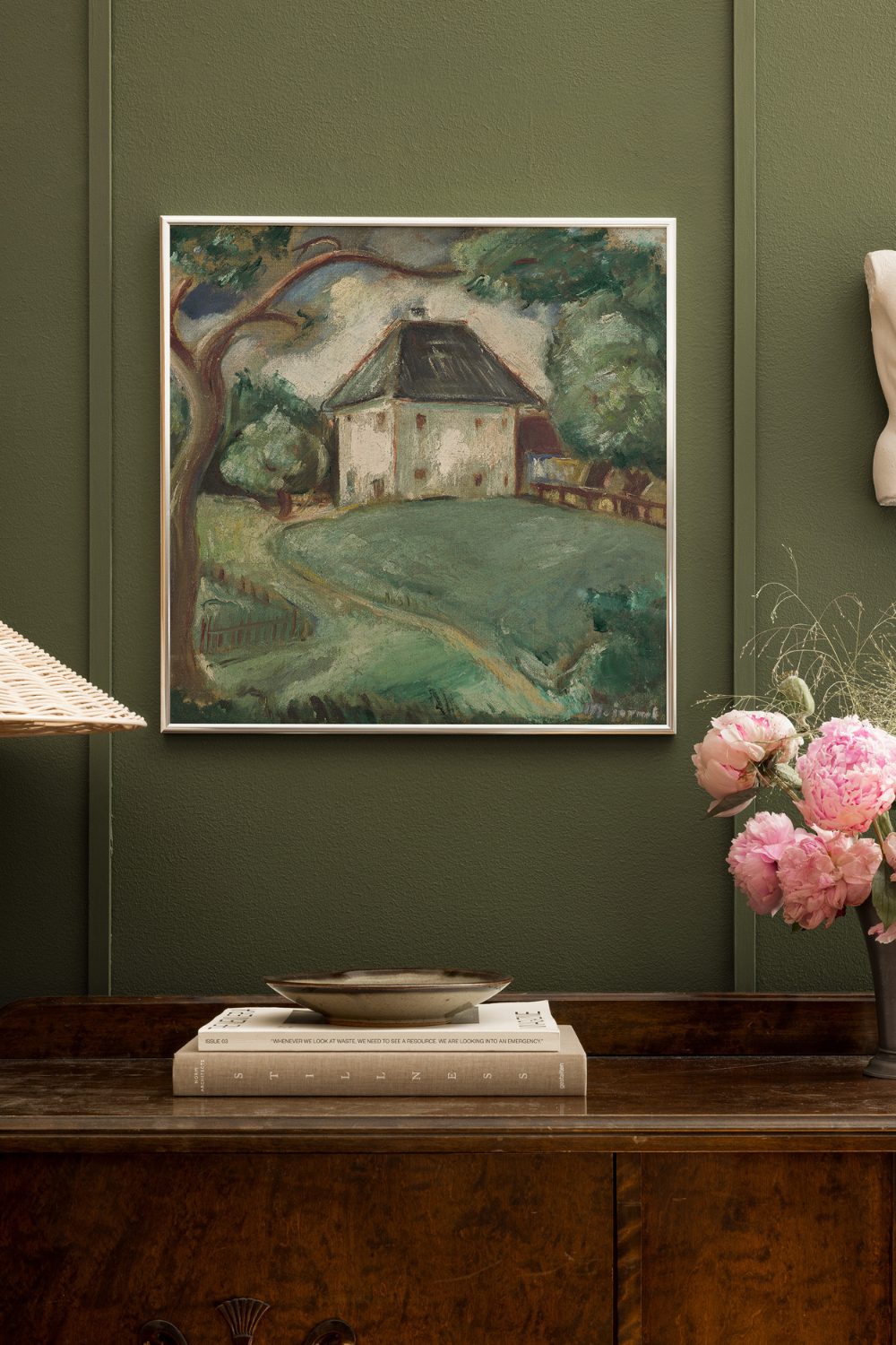 Painting of house and tree on an olive green wall above a wooden shelf holding books and vase of pink flowers