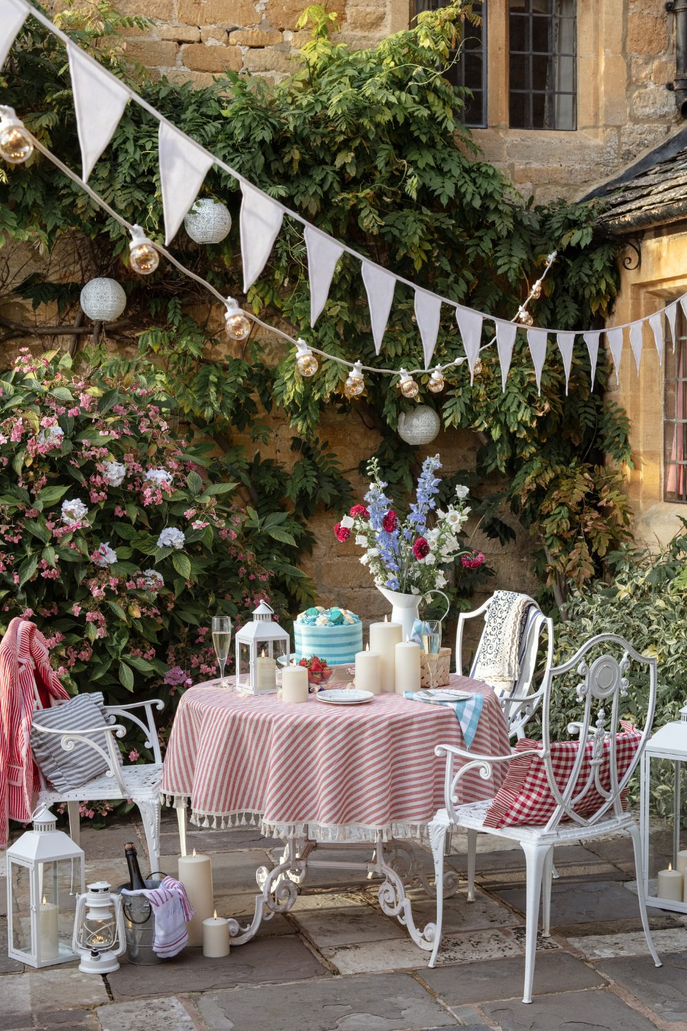 White metal chairs and table with striped tablecloth holding candles and vase of flowers on patio under white bunting