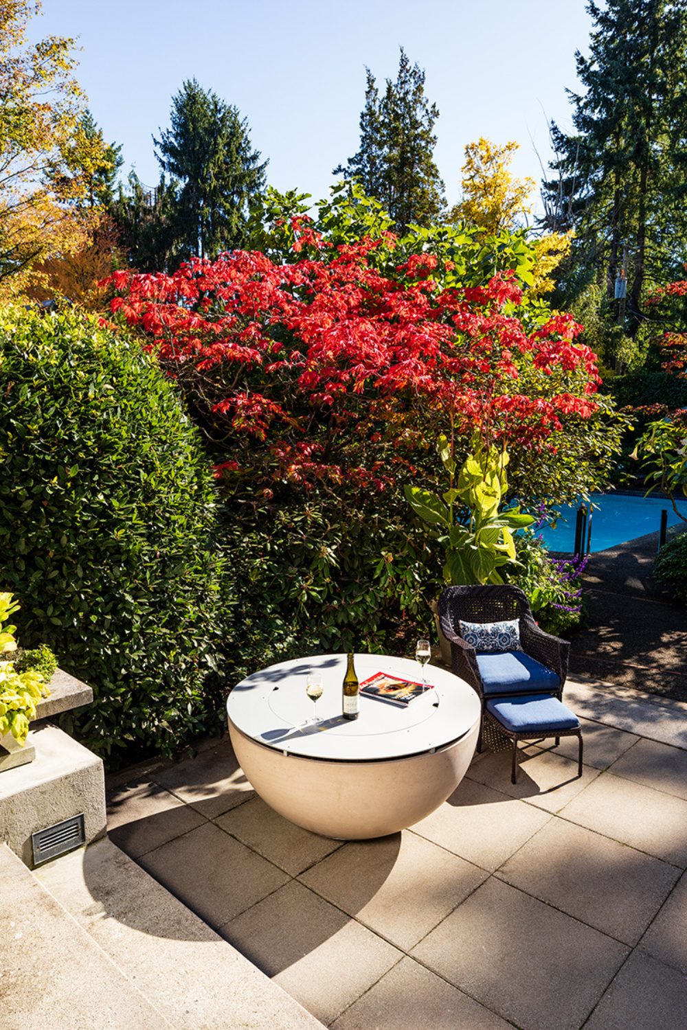 Light grey patio with round fire pit and wicker chair in front of colourful trees