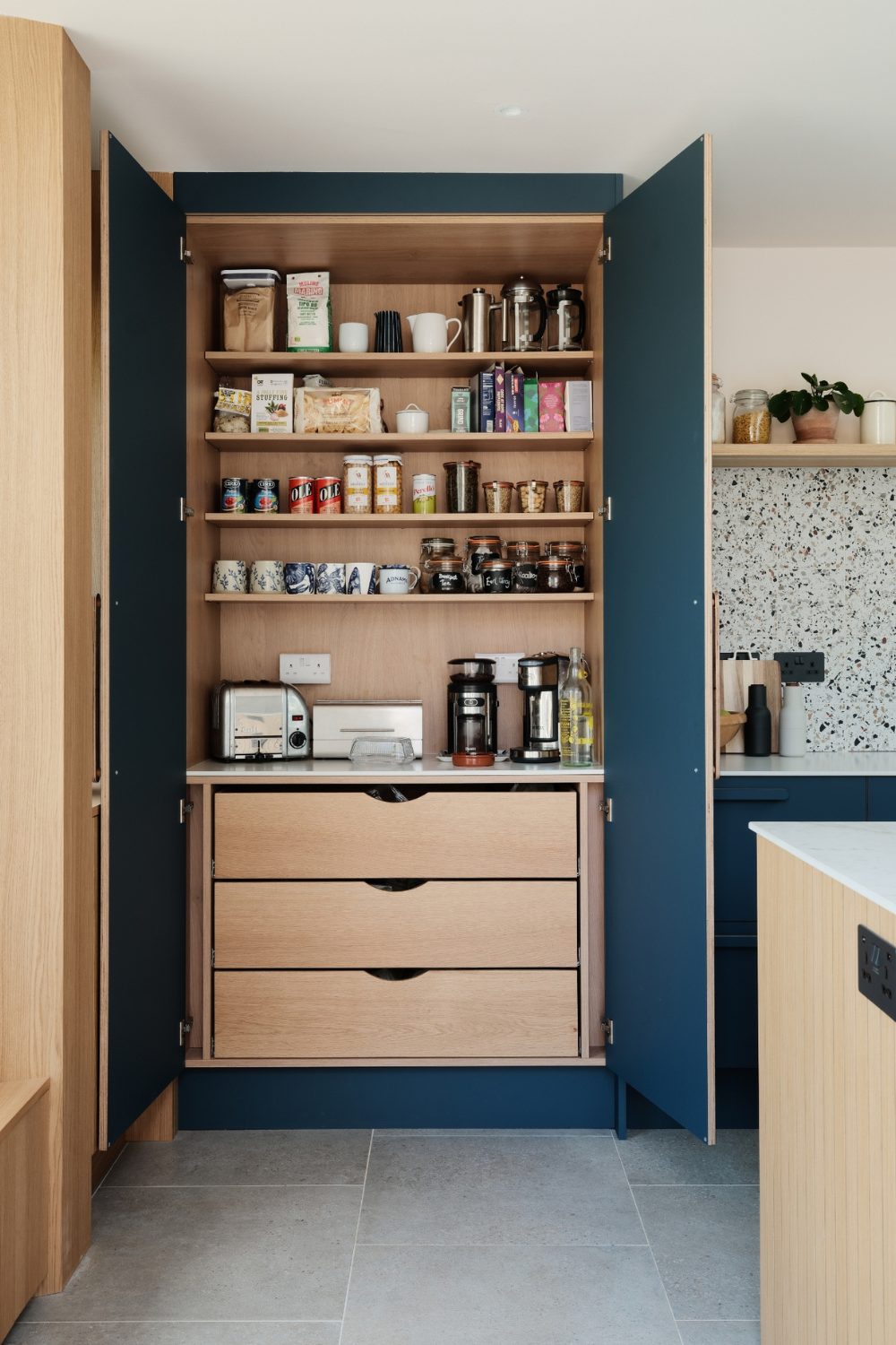 Blue and light wood kitchen cabinet with built-in shelving holding jars, cups and packets