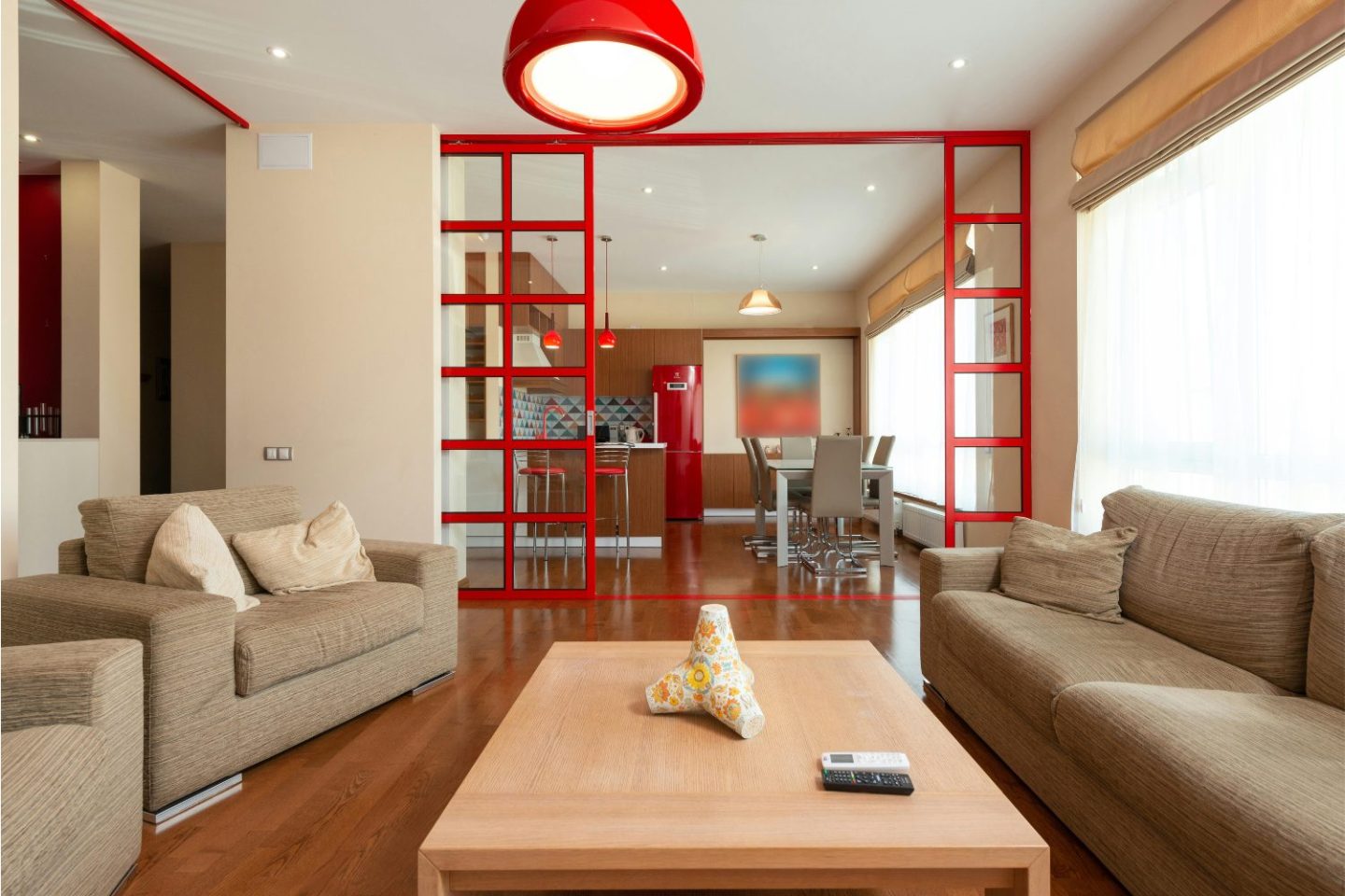 Glass and red steel door dividing a kitchen with red fridge and living room with brown sofas and armchairs, wooden coffee table and red light