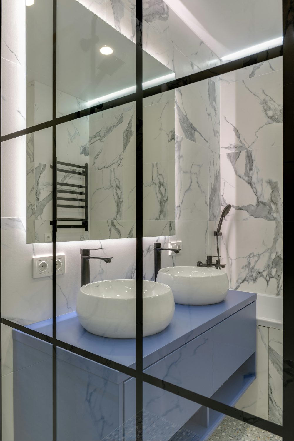 Glass and black steel door panes in front of a white marble bathroom with two white sinks on a blue cabinet