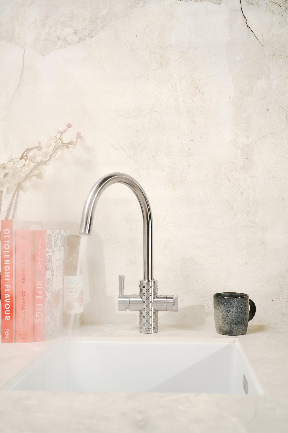 Silver tap with cross design next to grey mug and red cookbooks against a stone wall