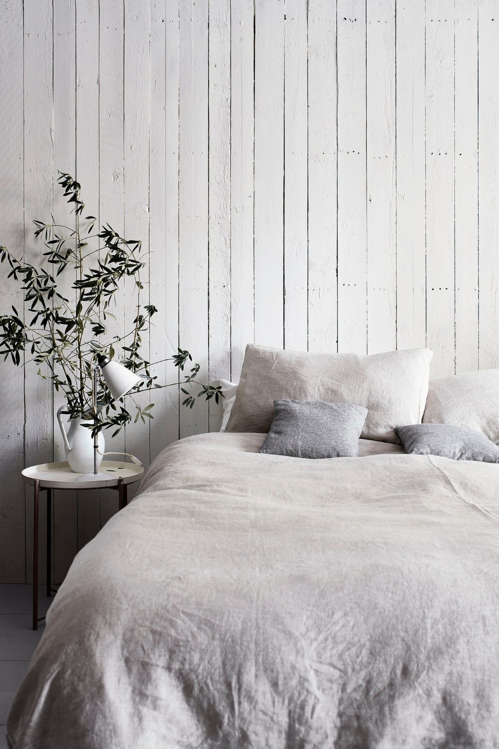 Natural finished white wooden wall behind white linen bed and white vase holding plant