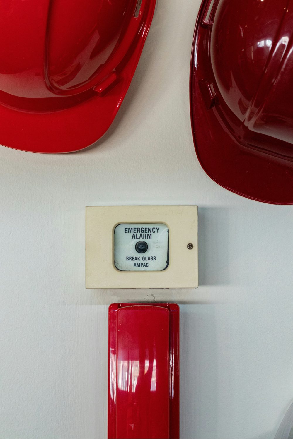 Two red hard hats hanging on white wall above fire alarm and red phone
