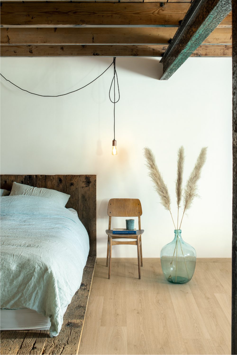 Light blue bed on rough wooden platform next to wooden chair and blue vase under exposed beams