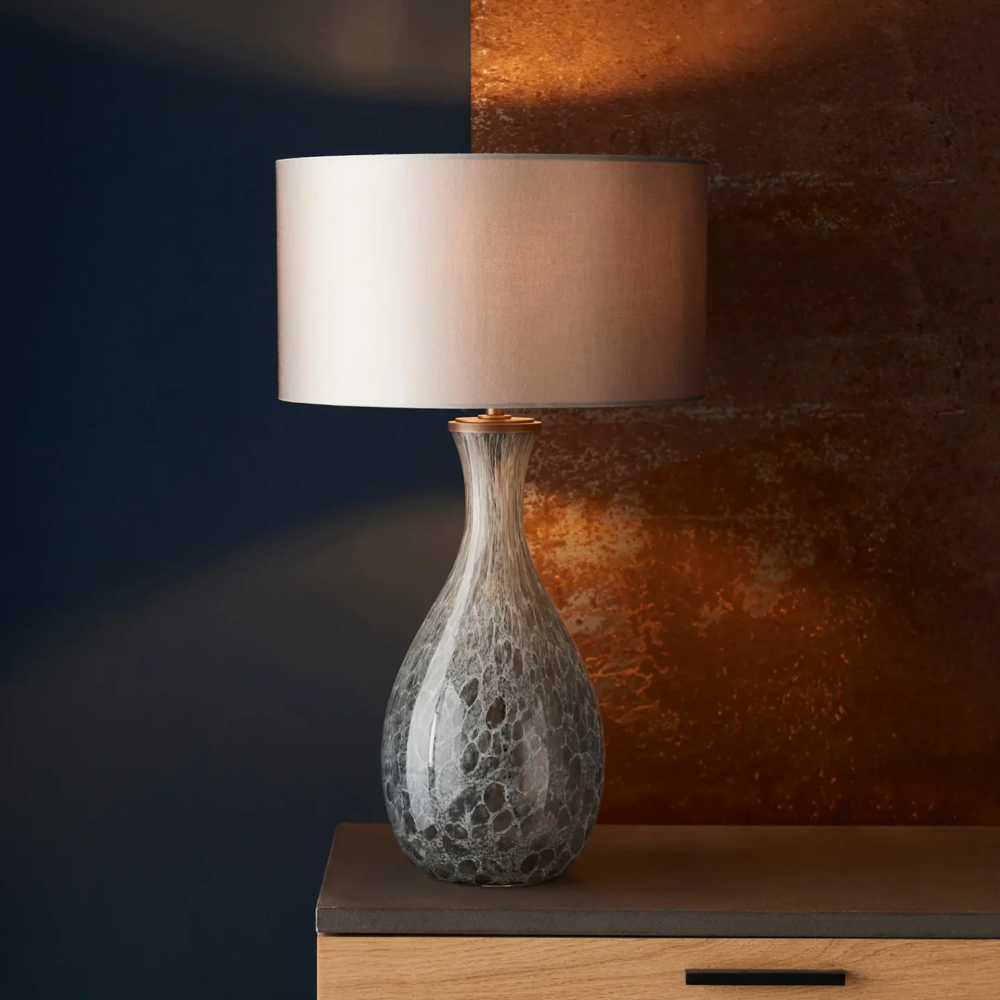 Lamp with grey glass base on wooden counter in front of orange and black wall