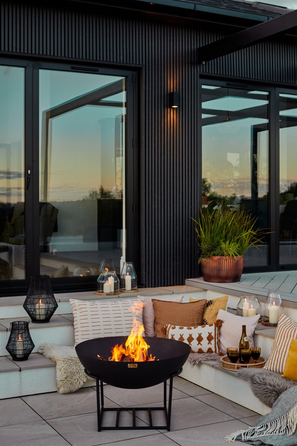 Fire pit with freestanding brazier surrounded by white steps with candles, throws and cushions in front of black and glass house
