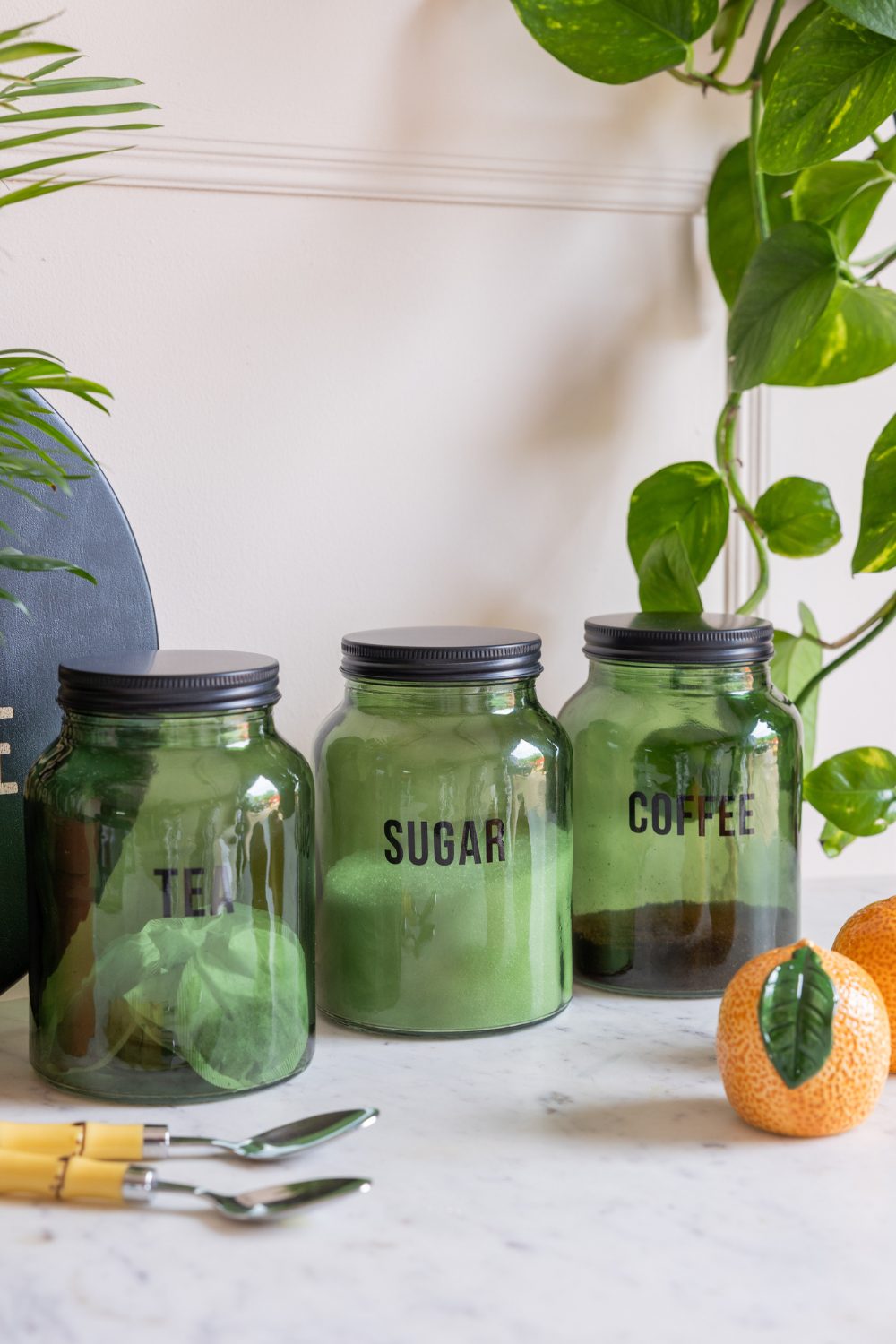 Three green glass jars marked Tea, Sugar and Coffee on white counter next to ceramic orange, spoons and plants