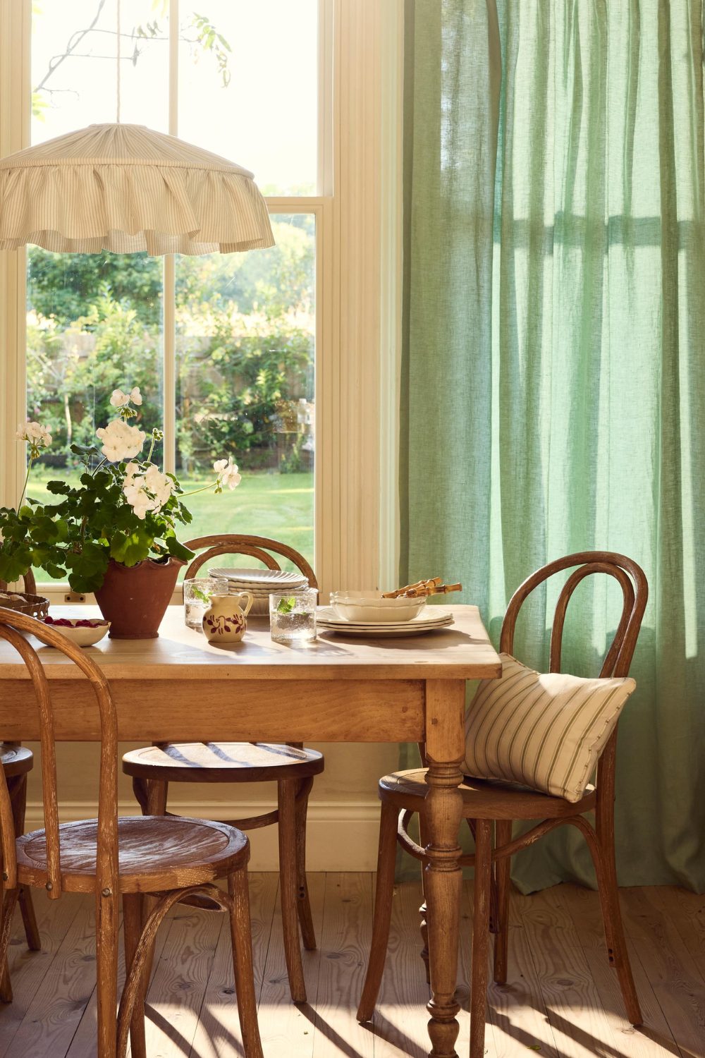 White kitchen with green linen curtains, wooden table and chairs in front of window with garden view