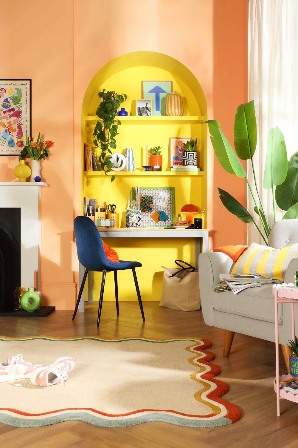 Yellow alcove in orange living room with built-in shelves holding plant pots, stationery and lamp, next to grey armchair and blue upright chair on wooden floor with white rug