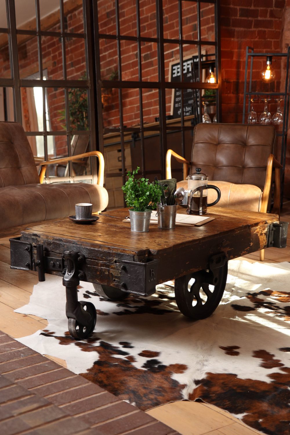 Brick living room with mirrored back wall, leather armchairs, thick wooden table with metal wheels and a cowskin rug