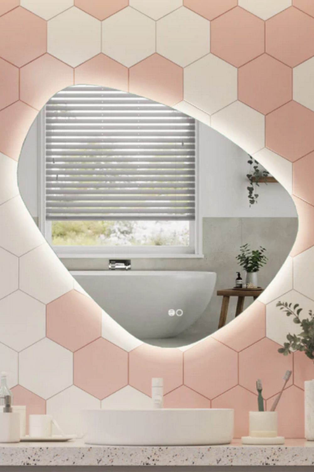 Irregularly-shaped mirror on pink and white tiled wall reflecting white bath in front of window with blinds