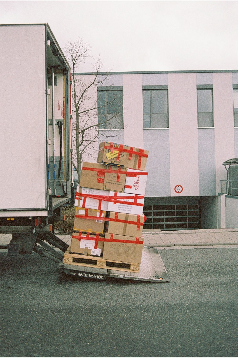Pallet of cardboard boxes secured with red tape being loaded into white truck with white building in background