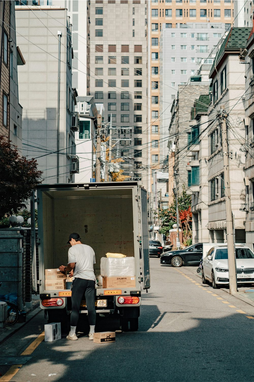 Man loads boxes into back of white truck in city street with telephone pole and high-rise buildings