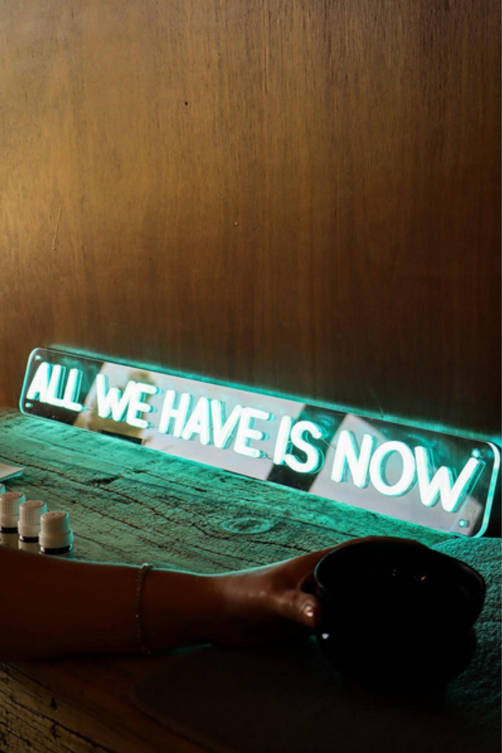 Blue neon 'All We Have Is Now' sign in front of wooden counter with hand holding cup