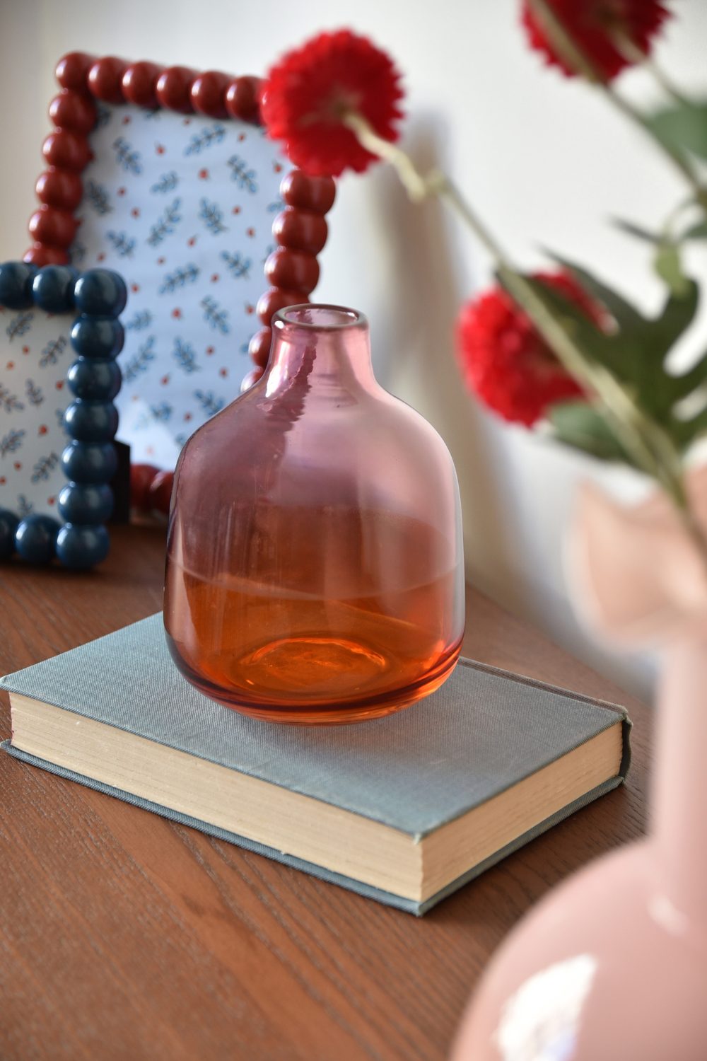 Red glass vase on blue book in front of red flowers and photo frame