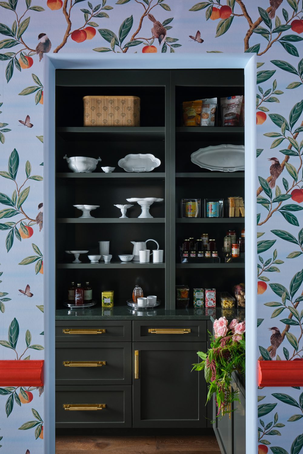 Dark shelves holding white cake stands and crockey in blue orange-patterned wallpaper with red wood detailing