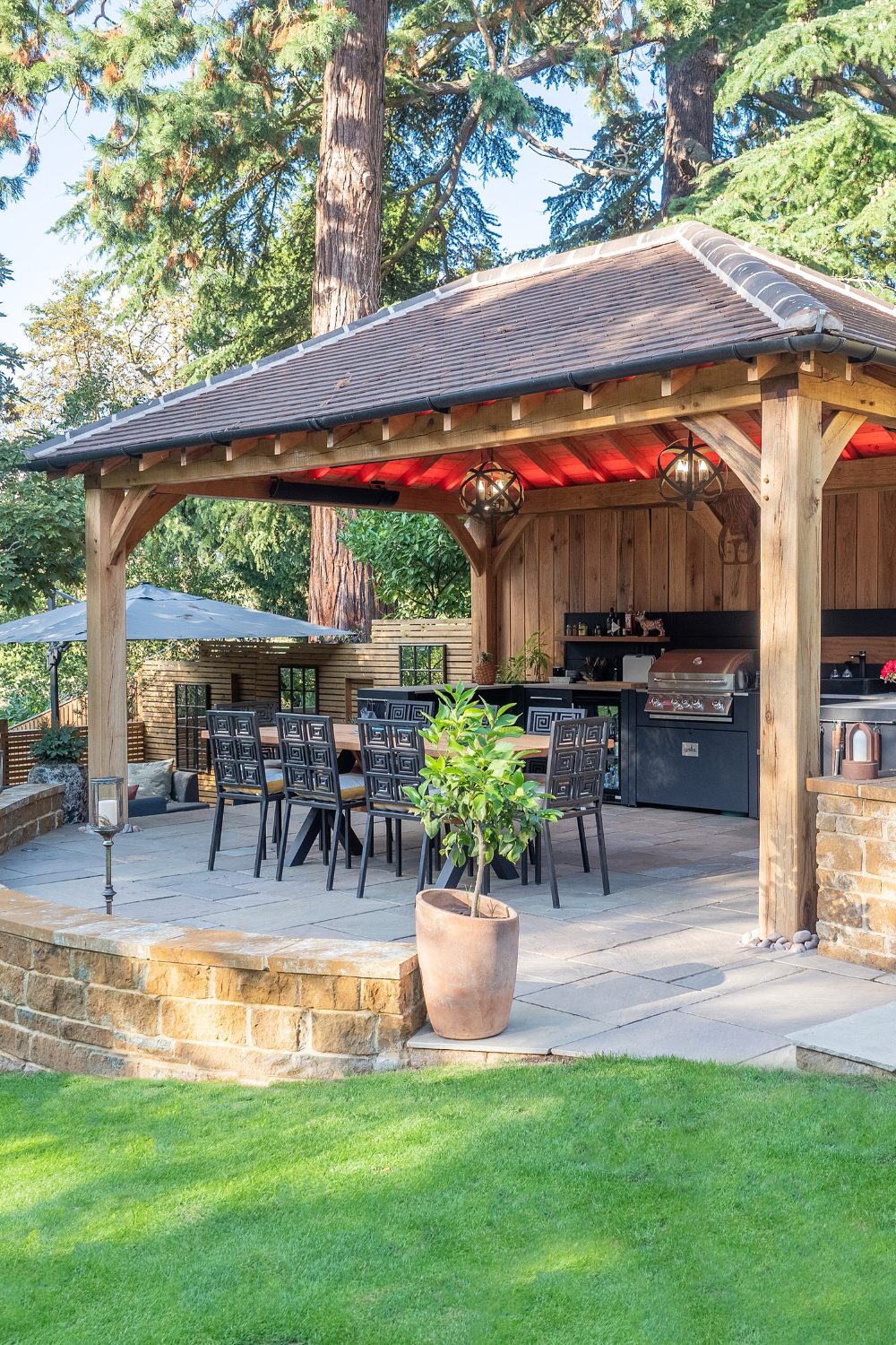 Garden wooden pergola over wooden table, metal chairs and potted plants behind a green lawn
