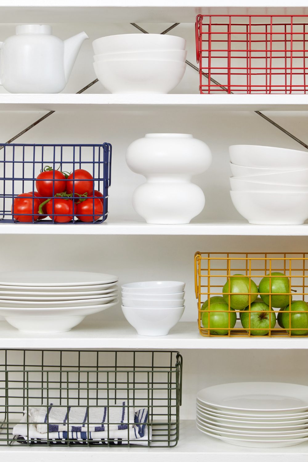 White shelves holding wire baskets of tomatoes and apples, and stacks of white bowls,