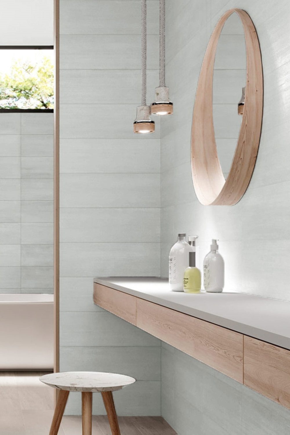 White tiled bathroom with wooden counter holding cosmetic bottles, wooden stool, round mirror and hanging lamps