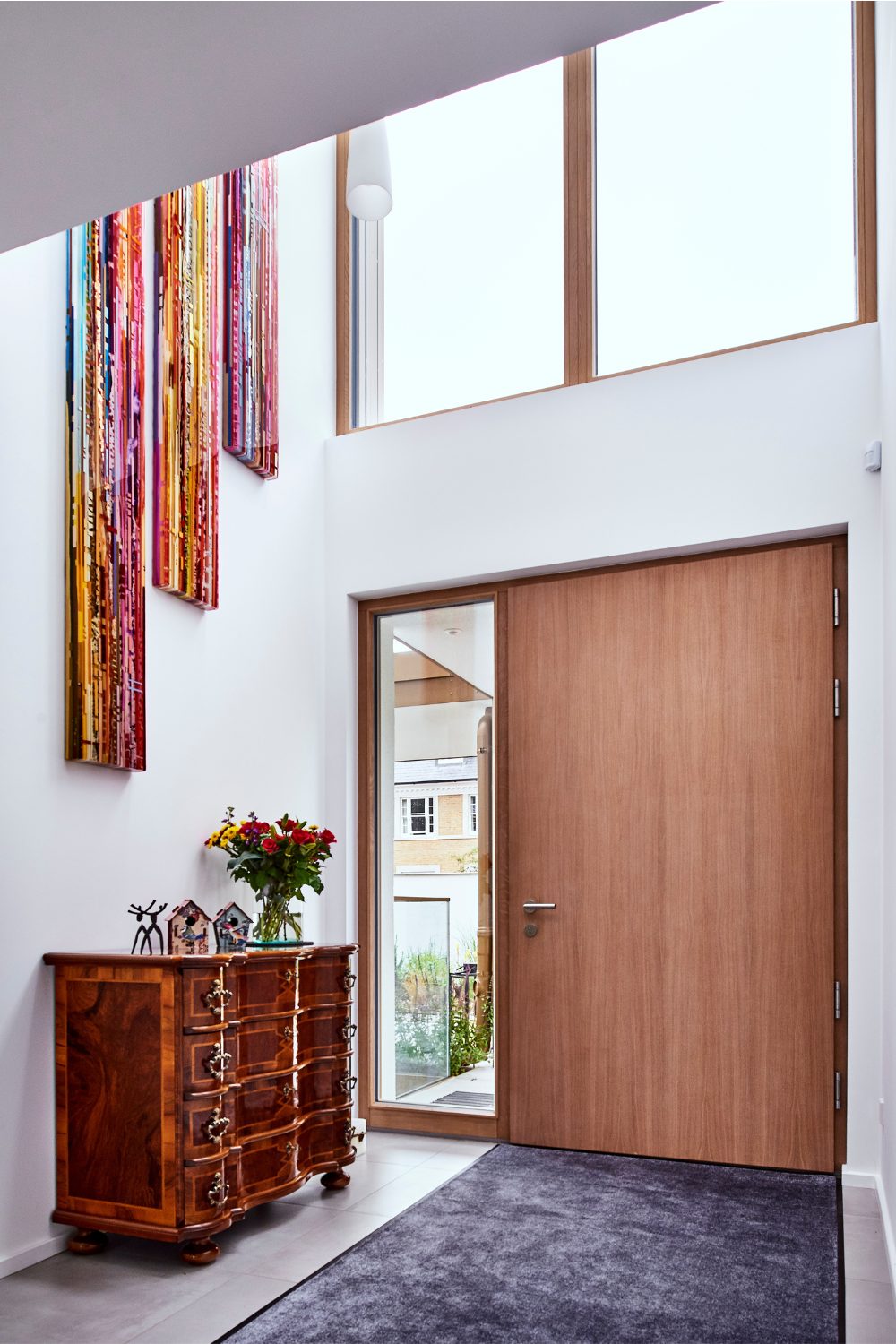 Double-storey white entrance hall with wooden front door, wooden chest of drawers, and colourful vertical wall art