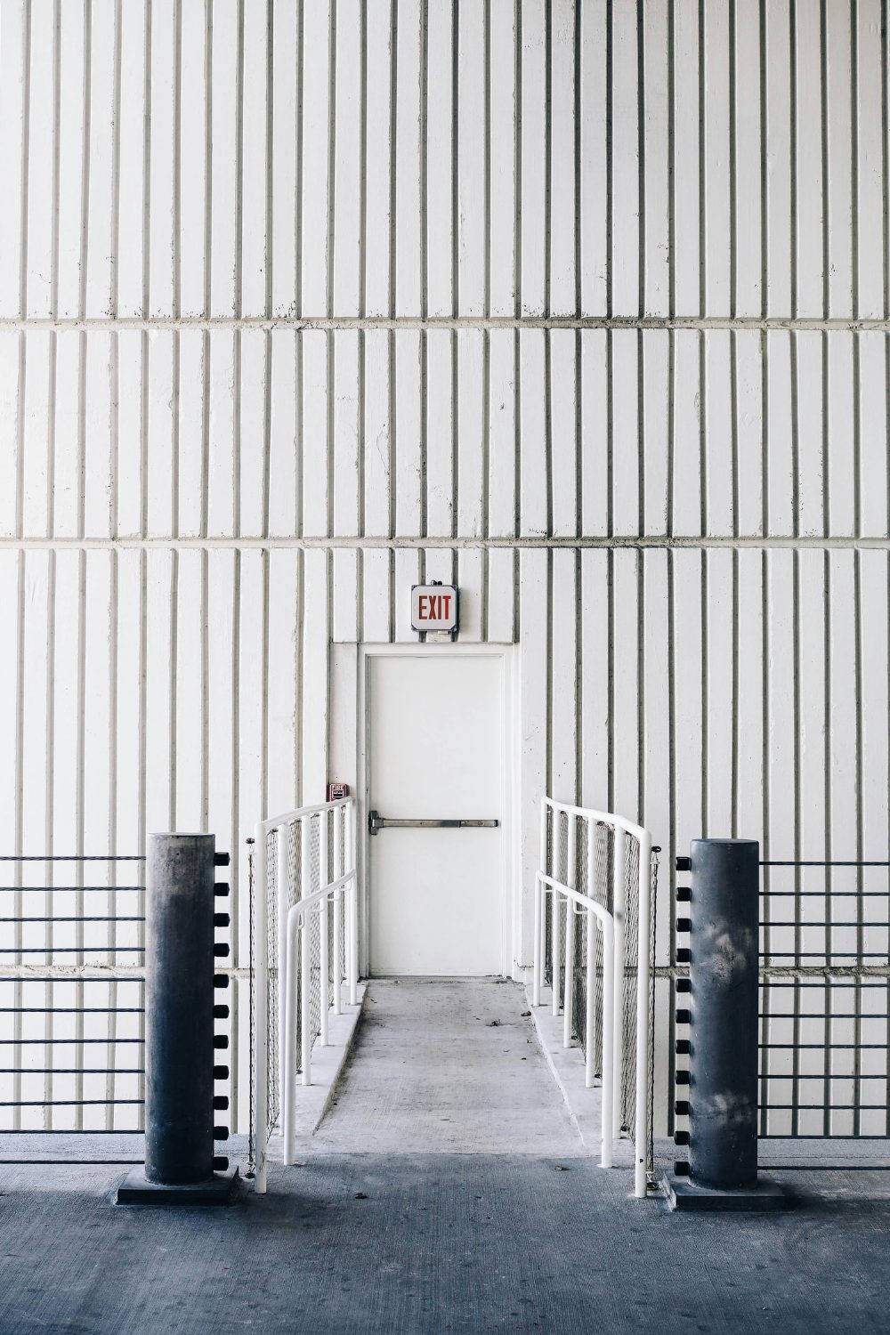 White outer door in white tiled wall at end of concrete bridge lined by white metal railings