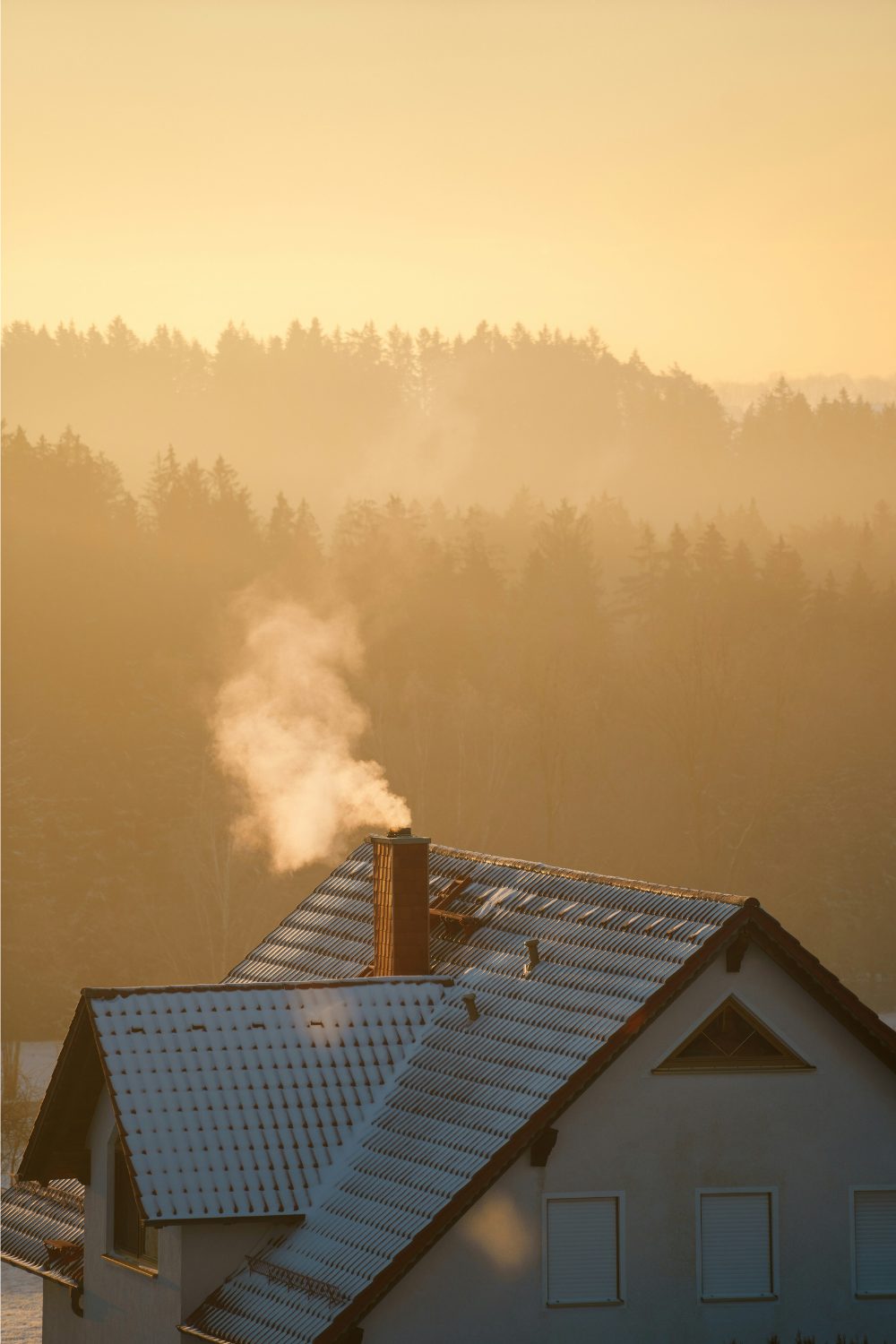 Snow-covered house roof with steam coming from chimney in front of forest