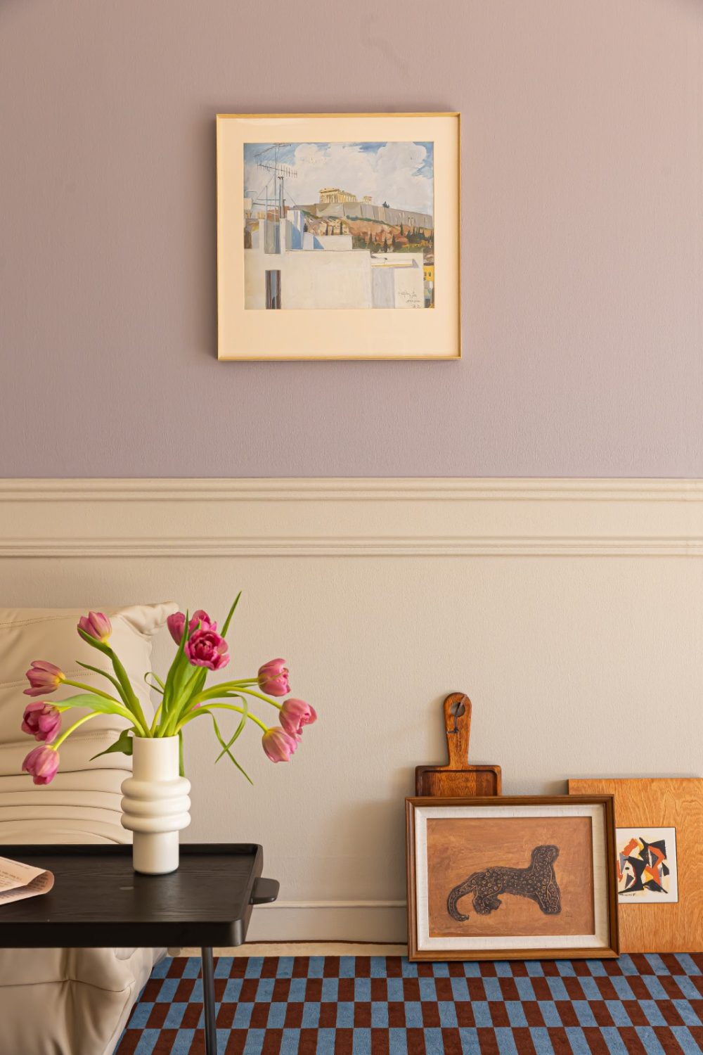 Framed prints propped against dusky pink wall with white wood panelling next to white chair and black coffee table holding white vase of pink flowers