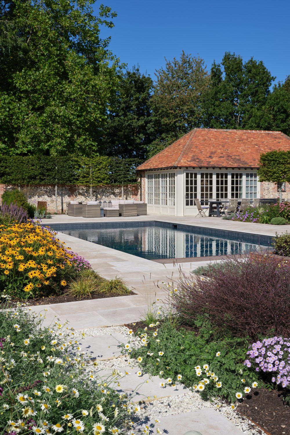 Pool with surrounding white stone and flowerbeds with colourful flowers, in front of brick poolhouse