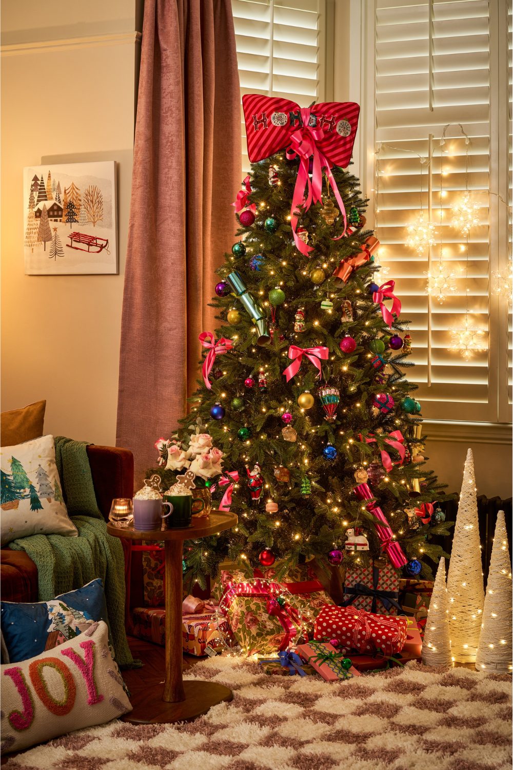 Christmas tree topped with red bow with presents at the bottom next to brown sofa with green throw, wooden table, and white decorative cones