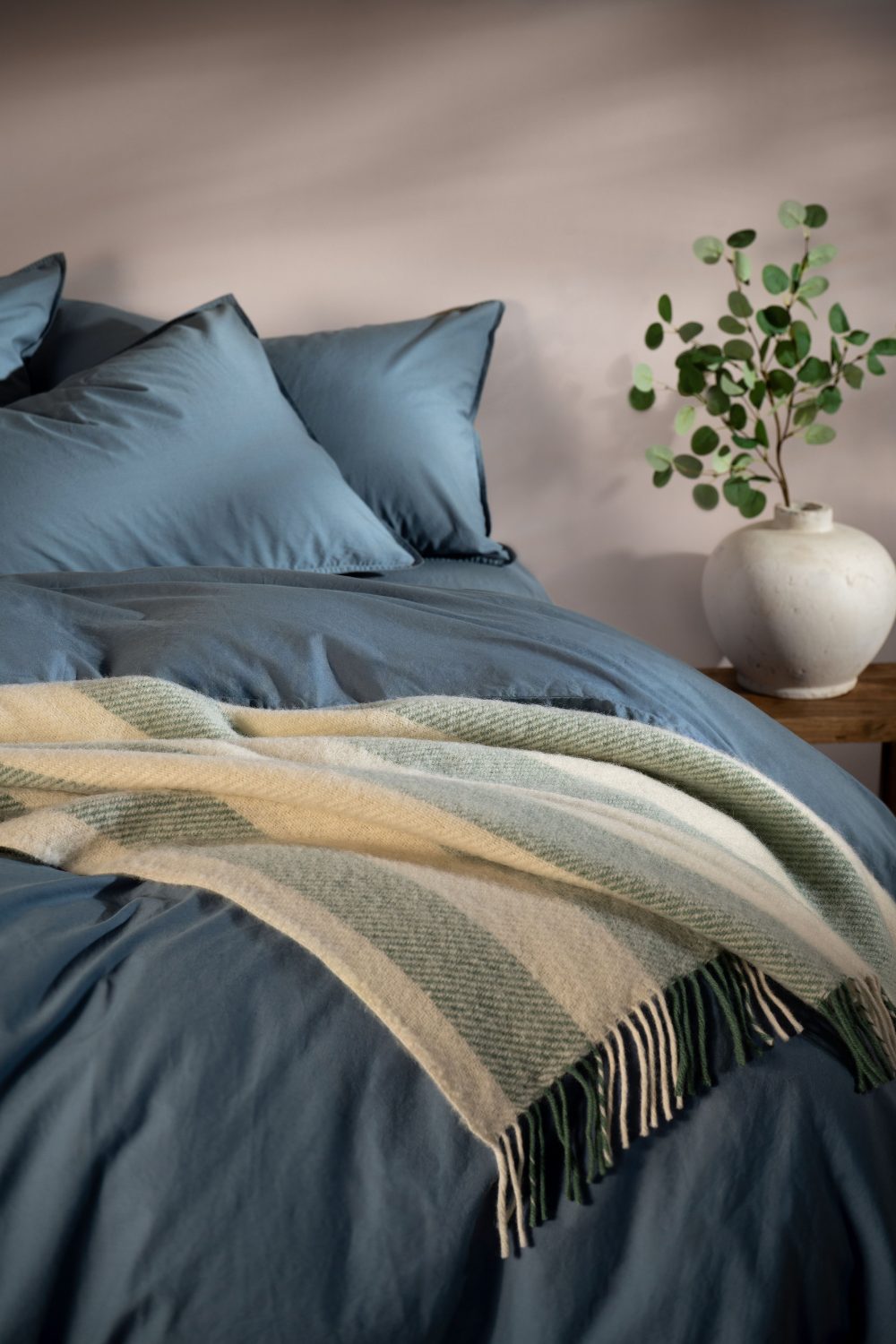 Blue bed with white and green wool throw next to a wooden table holding white vase