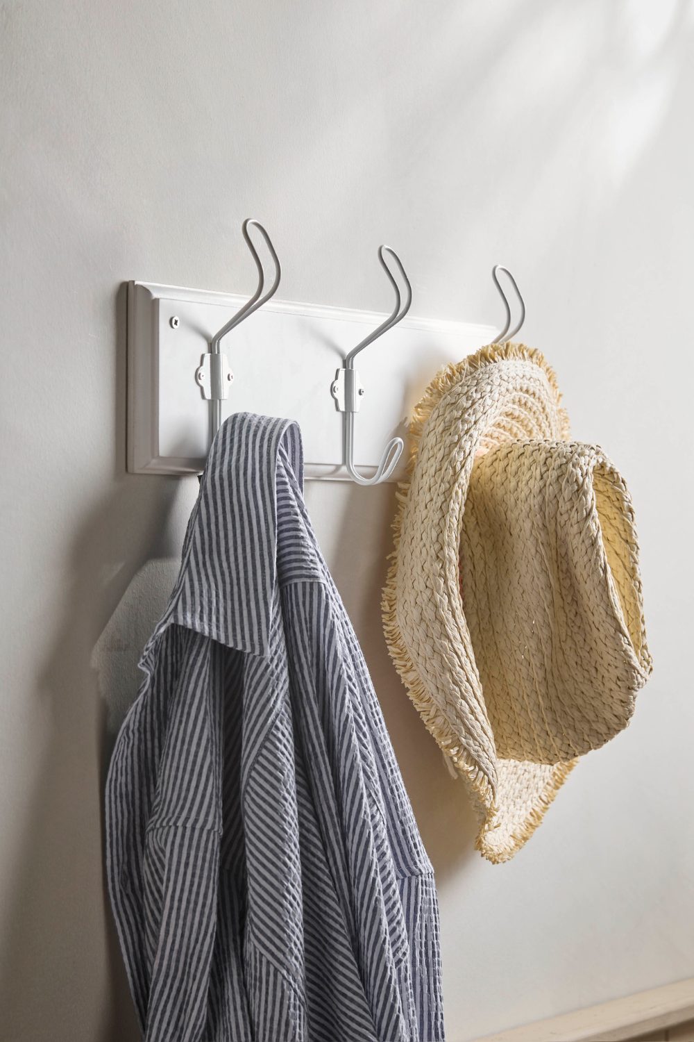 White metal coat hook on white wall holding straw hat and blue striped shirt