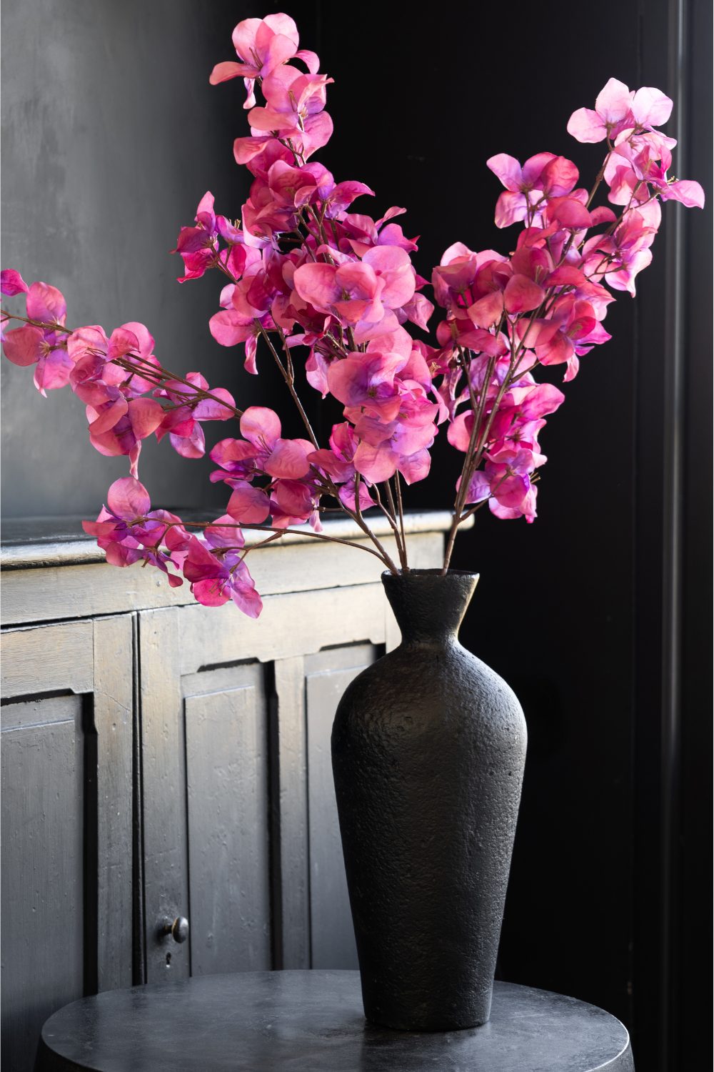 Vibrant pink flowers in a black vase on round black table in front of black cabinet
