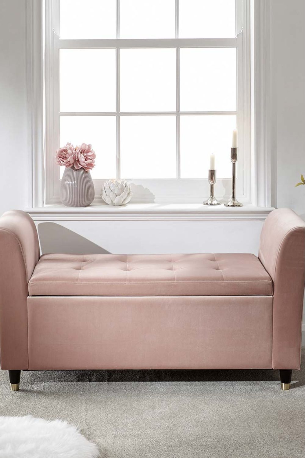 Pink window seat in front of white window with gold candlesticks and grey vase