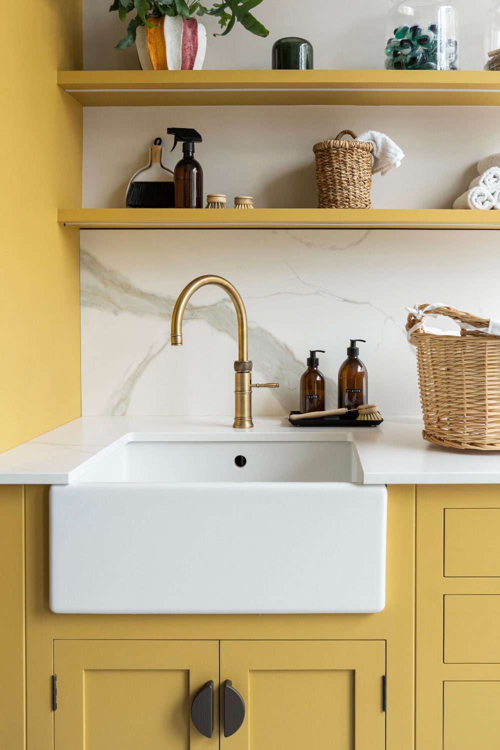 White kitchen sink with yellow cabinet and shelving holding vases, bottle and wicker basket