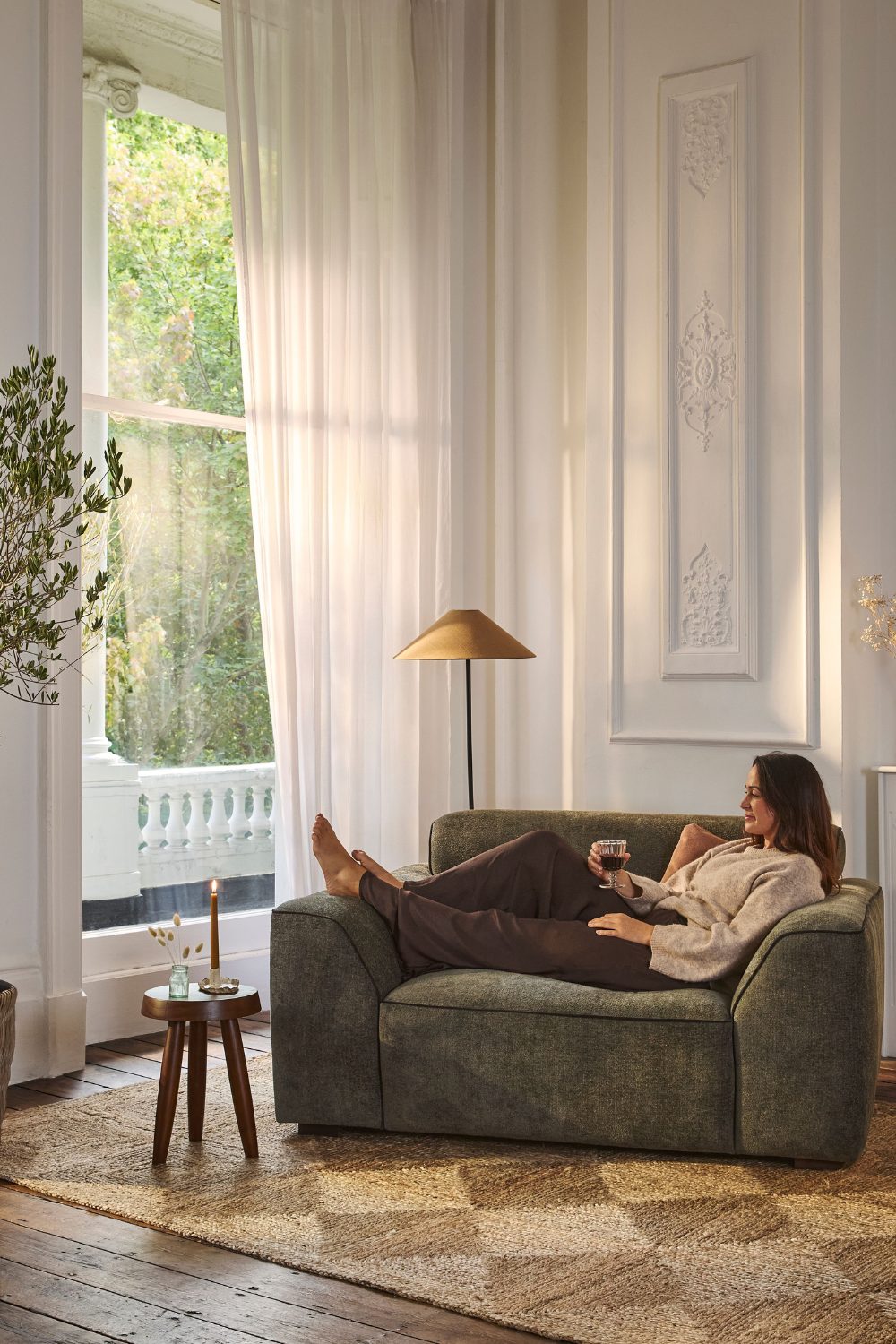 Woman sitting in dark green armchair on checkerboard jute rug by tall window with tree view
