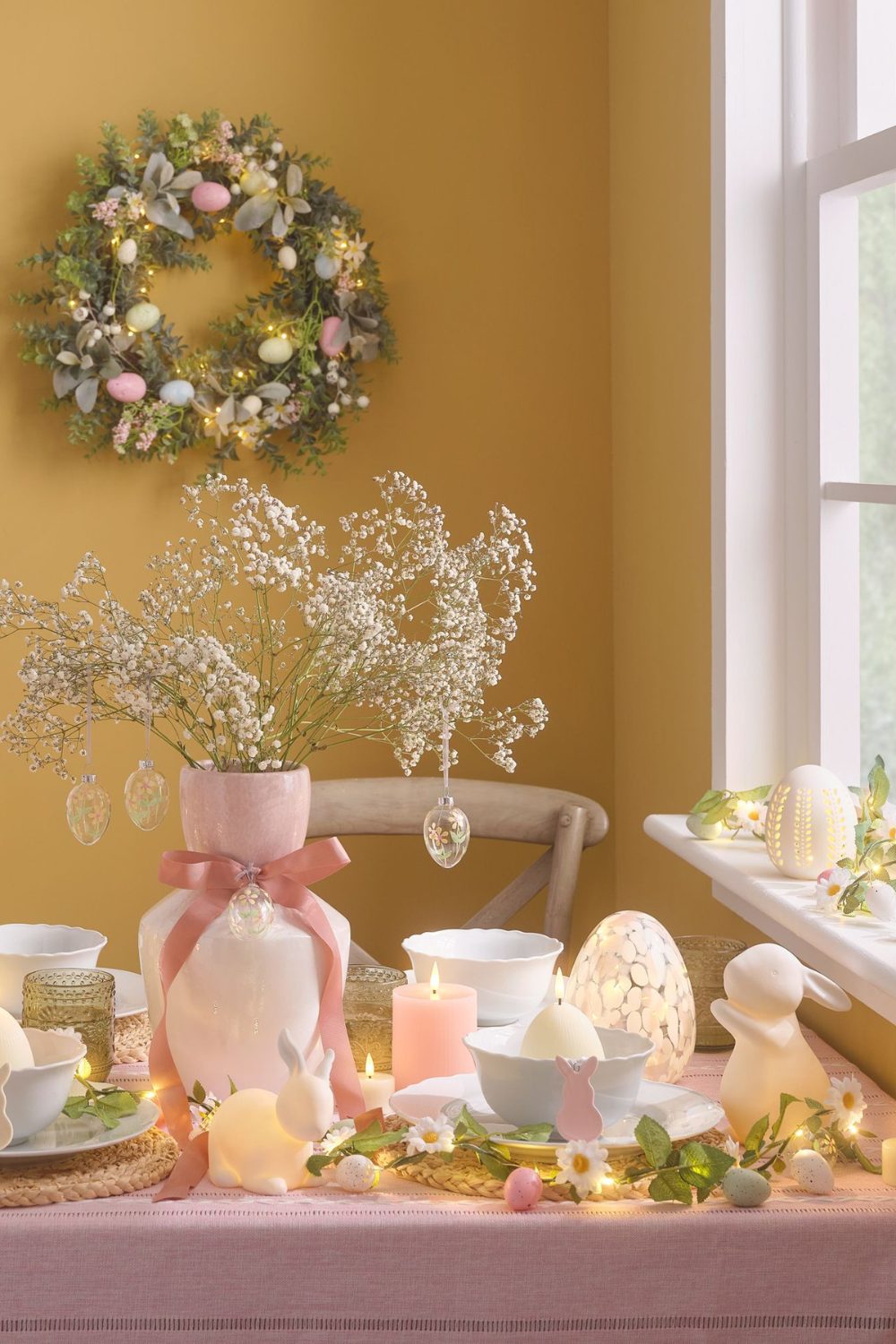 Table covered with pink tablecloth, with bunny lights, candles, and pink vase holding white flowers against yellow wall