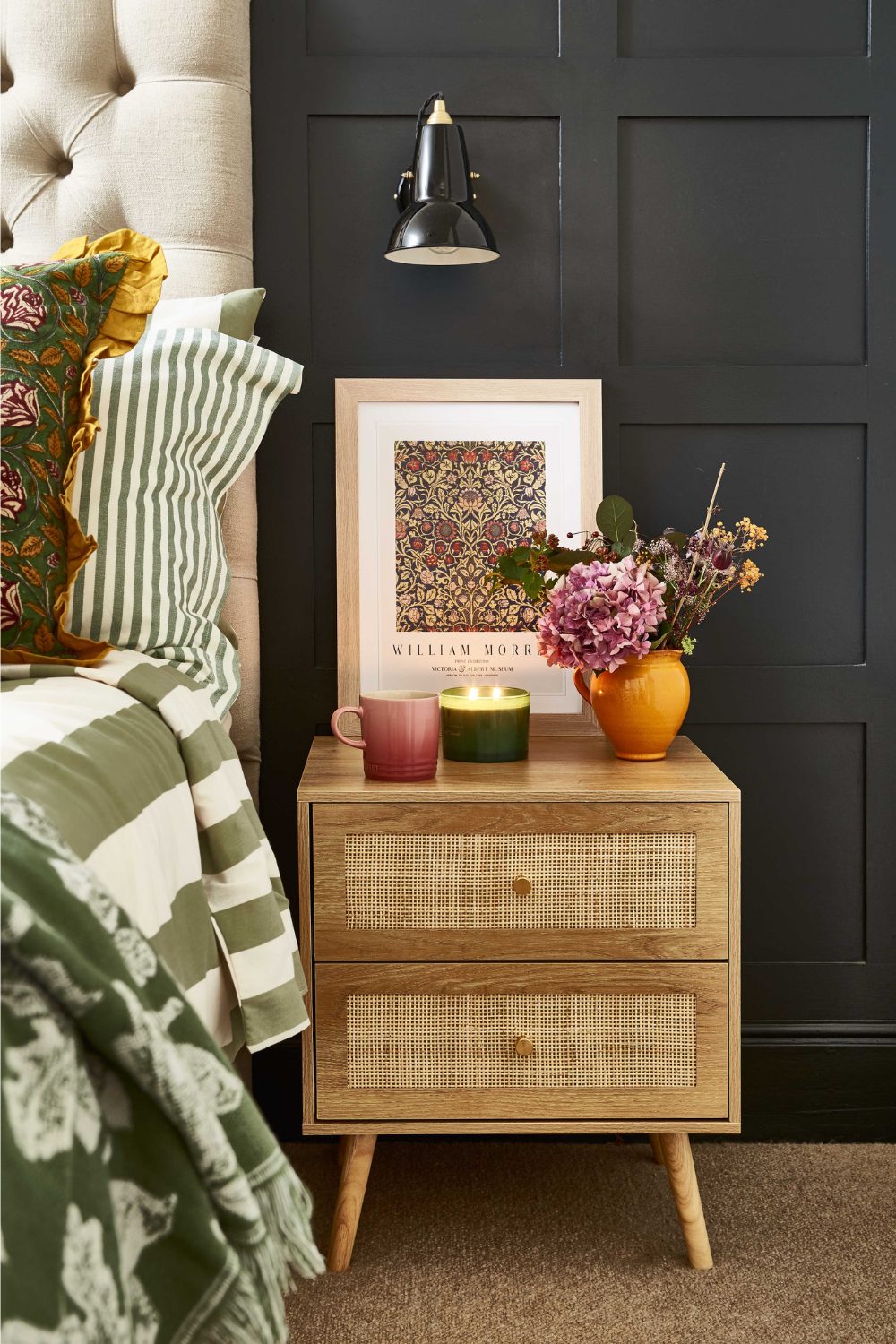 Wooden bedside table holding candle, mug and vase next to green bed in black wood-panelled bedroom