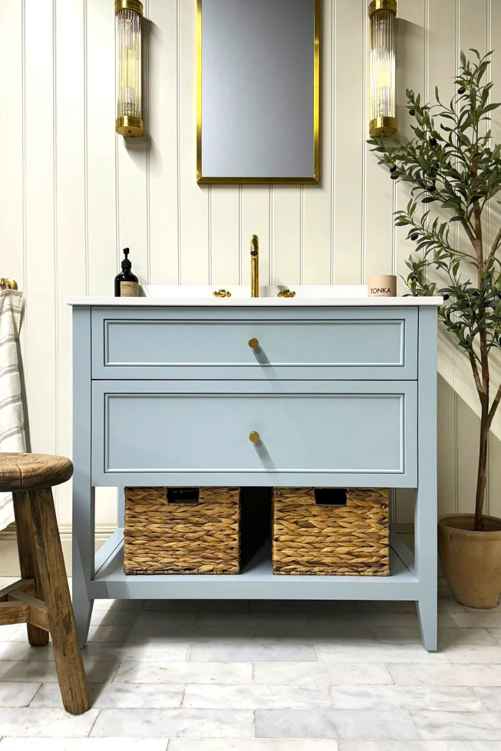 Potted tree and blue sink cabinet with woven storage baskets below rectangular mirror in cream wooden-panelled bathroom