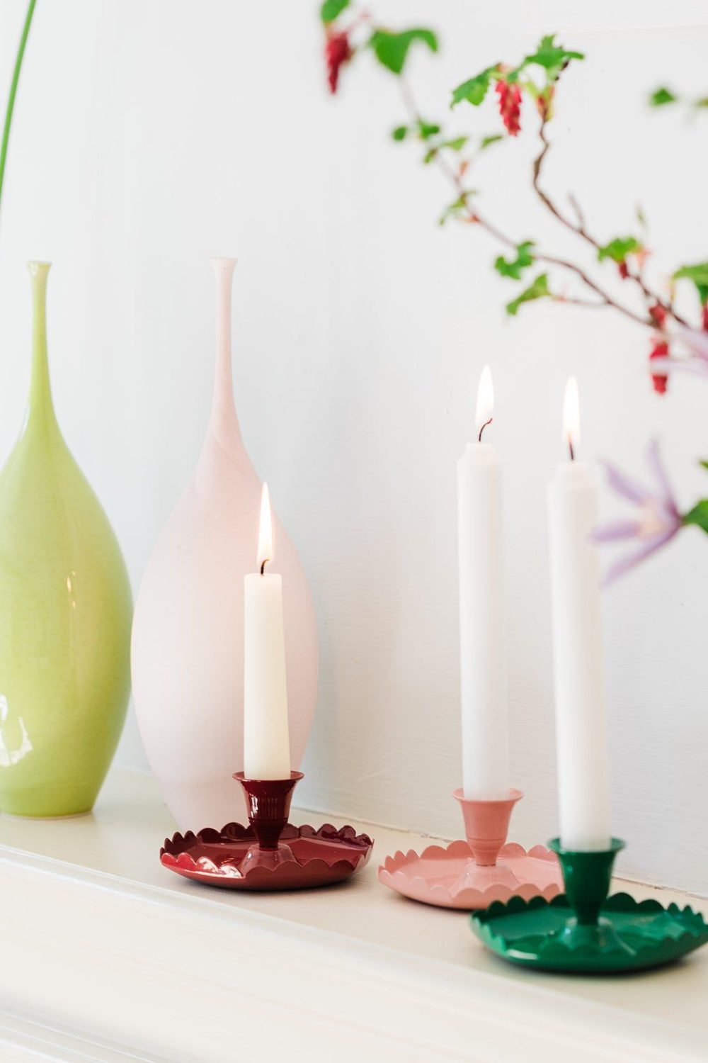 Three coloured candle holders with white candles, with pink and green vases in background