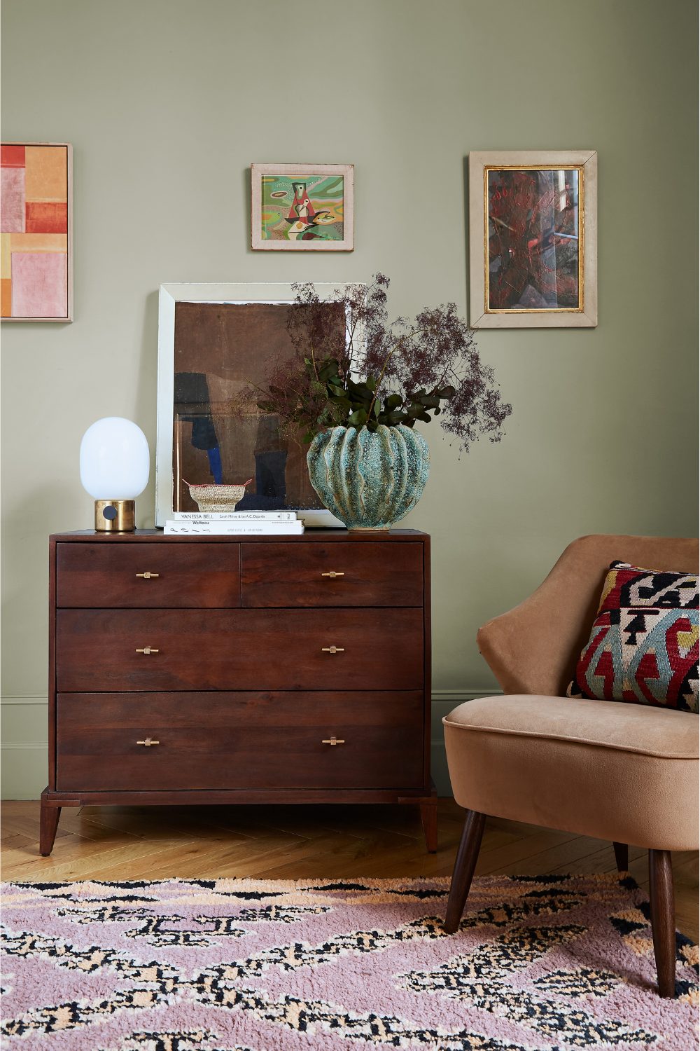 Wooden chest of drawers holding vases, lamp and framed art print, next to brown chair on pink patterned rug in muted green living room