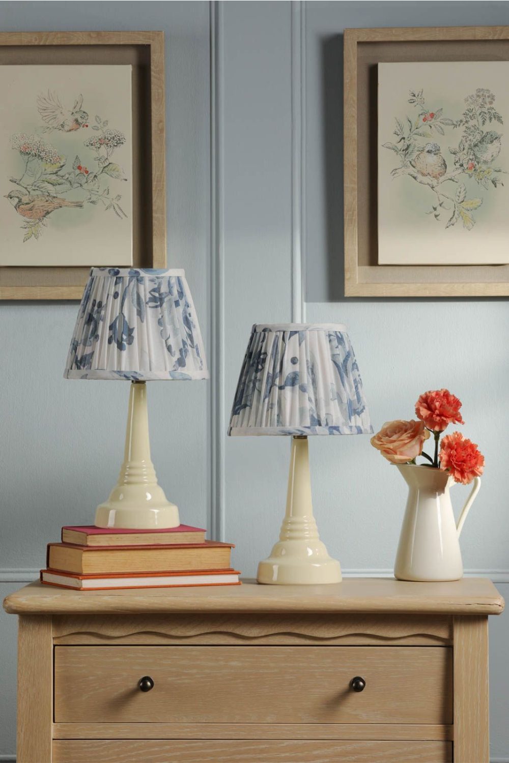 Wooden cabinet holding two blue and white floral lampshades, and white vase of red flowers against light blue wall