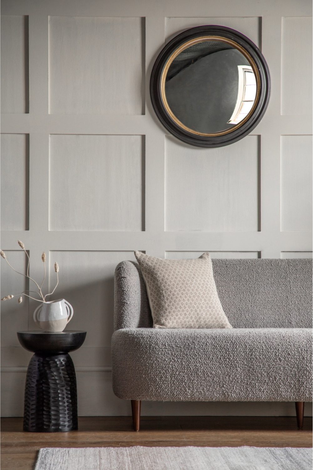 Grey living room with grey sofa, black side table holding brown vase, and grey rug below round mirror