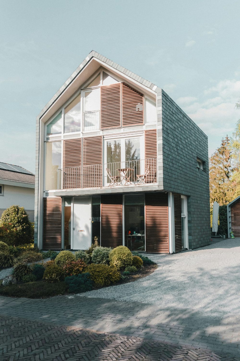 Three storey house with wooden panelling, balcony, and grey tiles with gravel drive and shrubs in front