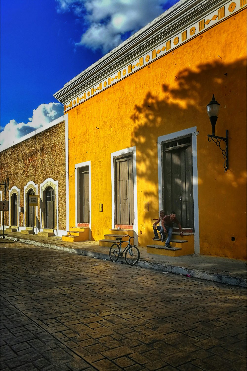 Yellow house with three wooden doors. Two men sit on the steps near a black bicycle.
